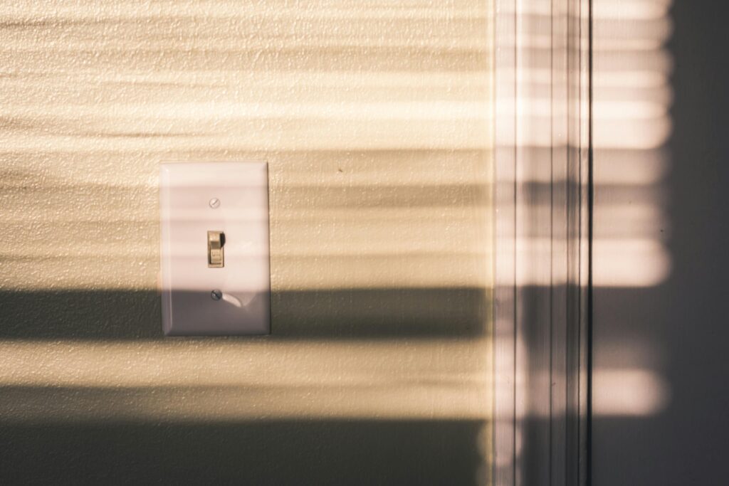 A light switch on a wall with dramatic shadows from the blinds in warm indoor lighting.